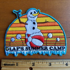 May include: A blue and orange embroidered patch with a snowman holding a red and white inner tube. The snowman is in front of a yellow, orange, and red sunset. The text "Olaf's Summer Camp" is at the bottom of the patch.