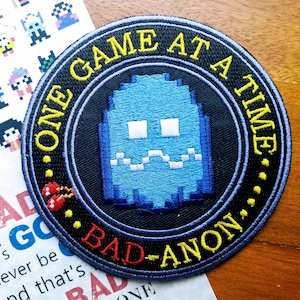 May include: A black and blue embroidered patch with a pixelated ghost and the text "ONE GAME AT A TIME BAD-ANON."