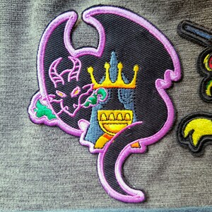 May include: A black and purple embroidered patch of a dragon wearing a gold crown. The dragon is curled around a golden goblet.