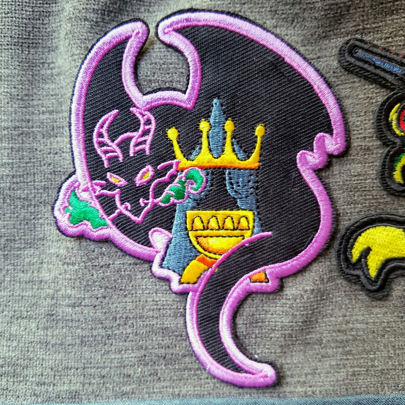 May include: A black and purple embroidered patch of a dragon wearing a gold crown. The dragon is curled around a golden goblet.
