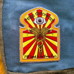May include: A yellow, red, and white embroidered patch with the text "Mara Optometry" and "We Look Into Your Eyes". The patch features a stylized eye and two snakes intertwined.
