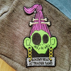 May include: Green and pink embroidered patch of a cartoon character with a skull-like face, wearing a pink hat with bone accents. The patch has the text "I LOST MY HEAD AT TRADER SAMS".