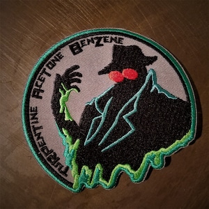 May include: Green, black, and red embroidered patch with a shadowy figure wearing a hat and the text "The Benzene Acetone"