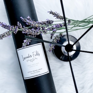 Op de afbeelding: Zwarte rietdiffuserfles met een wit etiket waarop staat "Lavender Fields English Lavender & Grey Sage Reed Diffuser Essential Oils Cotton Dreams London Scent of Deep Relaxation 100 ml / 3.38 fl oz". De fles is omgeven door lavendeltakken en zwarte diffuserstokjes.