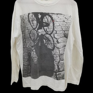 May include: White long-sleeve t-shirt with a black and white graphic print of a person riding a bicycle on a cobblestone street. The graphic print is centered on the shirt.