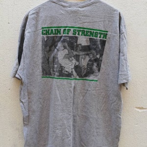 以下が含まれることがあります： 背面に人々の群衆の白黒写真がプリントされたグレーのTシャツ。写真の上に「CHAIN OF STRENGTH」という文字が緑色でプリントされています。