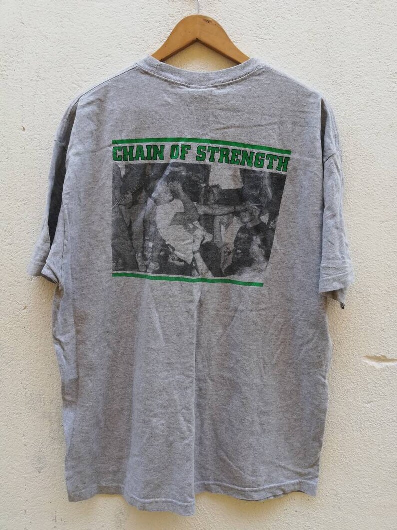 以下が含まれることがあります： 背面に人々の群衆の白黒写真がプリントされたグレーのTシャツ。写真の上に「CHAIN OF STRENGTH」という文字が緑色でプリントされています。