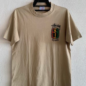 Puede incluir: Una camiseta de color beige con un gr&aacute;fico negro, verde, rojo y amarillo del logotipo de Stussy con el texto "IRIE" debajo.