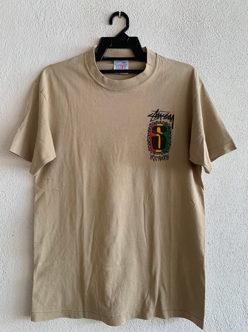 Puede incluir: Una camiseta de color beige con un gr&aacute;fico negro, verde, rojo y amarillo del logotipo de Stussy con el texto "IRIE" debajo.