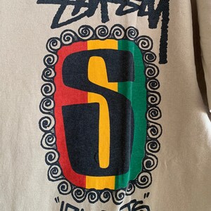 Puede incluir: Una camiseta beige con un logotipo "Stussy" en blanco y negro y una "S" negra en un c&iacute;rculo rojo, amarillo y verde con un borde decorativo. El texto "irie roots" est&aacute; escrito debajo del c&iacute;rculo.