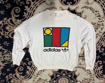 May include: White long-sleeve shirt with a colorful geometric design and the word "adidas" printed on the front.