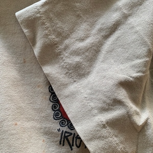 Puede incluir: Primer plano de una camiseta gris claro con un dise&ntilde;o gr&aacute;fico rojo y negro. El gr&aacute;fico presenta un c&iacute;rculo con un patr&oacute;n de espiral y la palabra "Riot" escrita debajo.