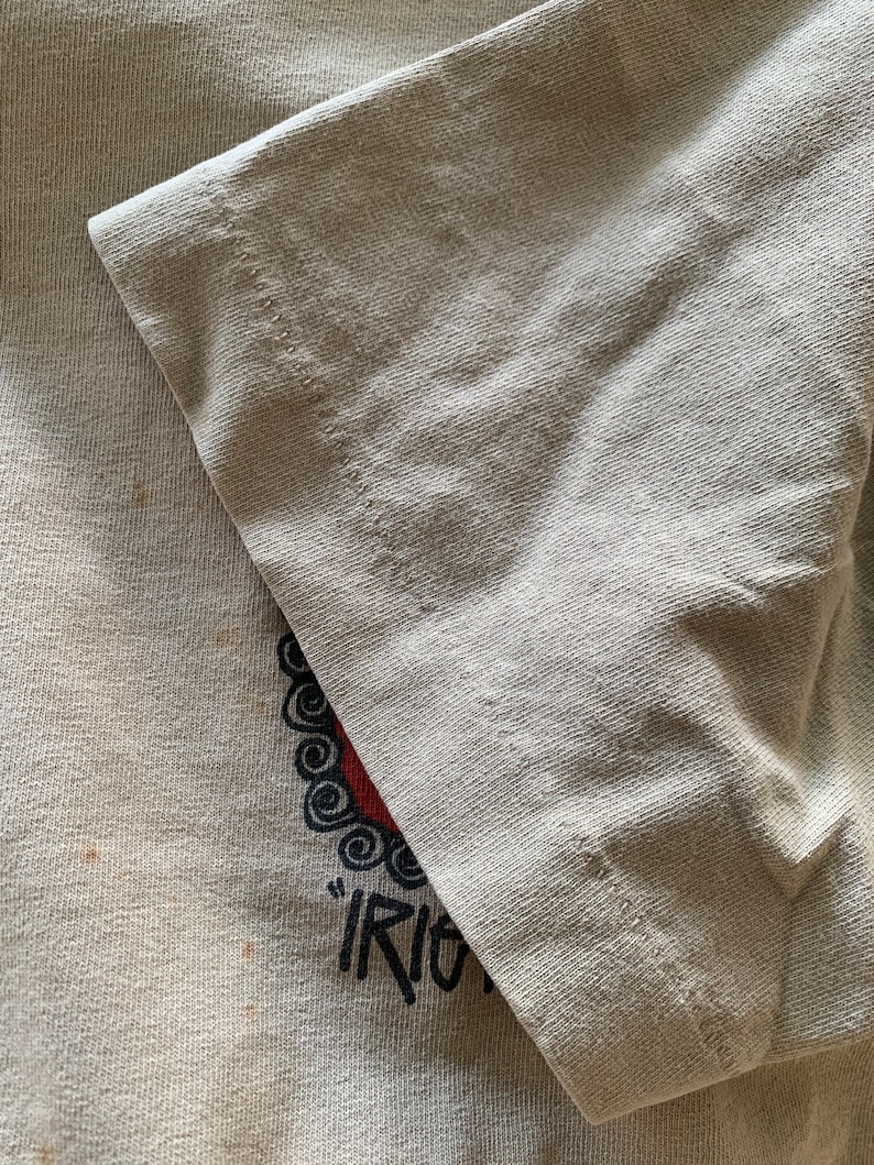 Puede incluir: Primer plano de una camiseta gris claro con un dise&ntilde;o gr&aacute;fico rojo y negro. El gr&aacute;fico presenta un c&iacute;rculo con un patr&oacute;n de espiral y la palabra "Riot" escrita debajo.