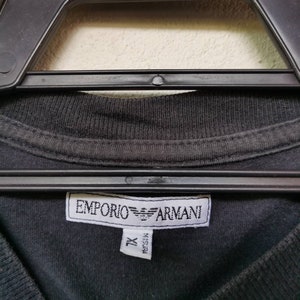 Puede incluir: Camiseta negra con una etiqueta blanca que dice "EMPORIO ARMANI" y "XL" en negro.