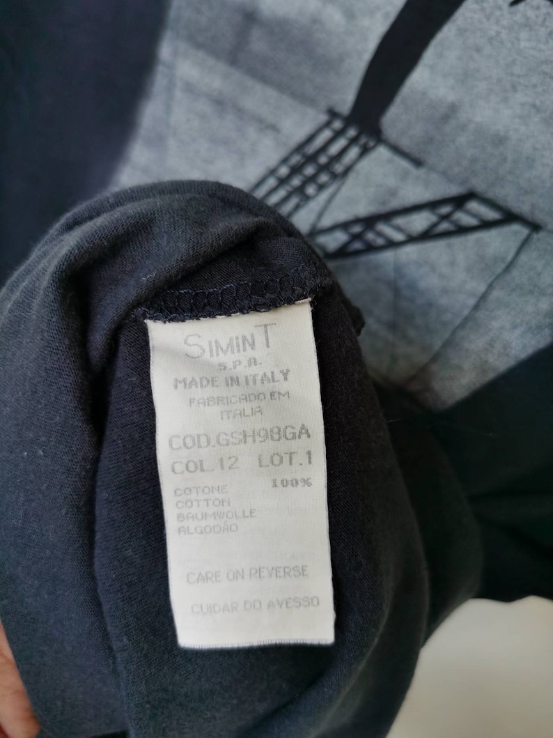 Puede incluir: Etiqueta blanca con texto negro en una camiseta negra. La etiqueta dice "SIMINT S.P.A. MADE IN ITALY FABRICADO EM ITALIA COD.GSH98GA COL.12 LOT.1 COTONE 100% COTTON BAUMWOLLE ALGOD&Atilde;O CARE ON REVERSE CUIDAR DO AVESSO"