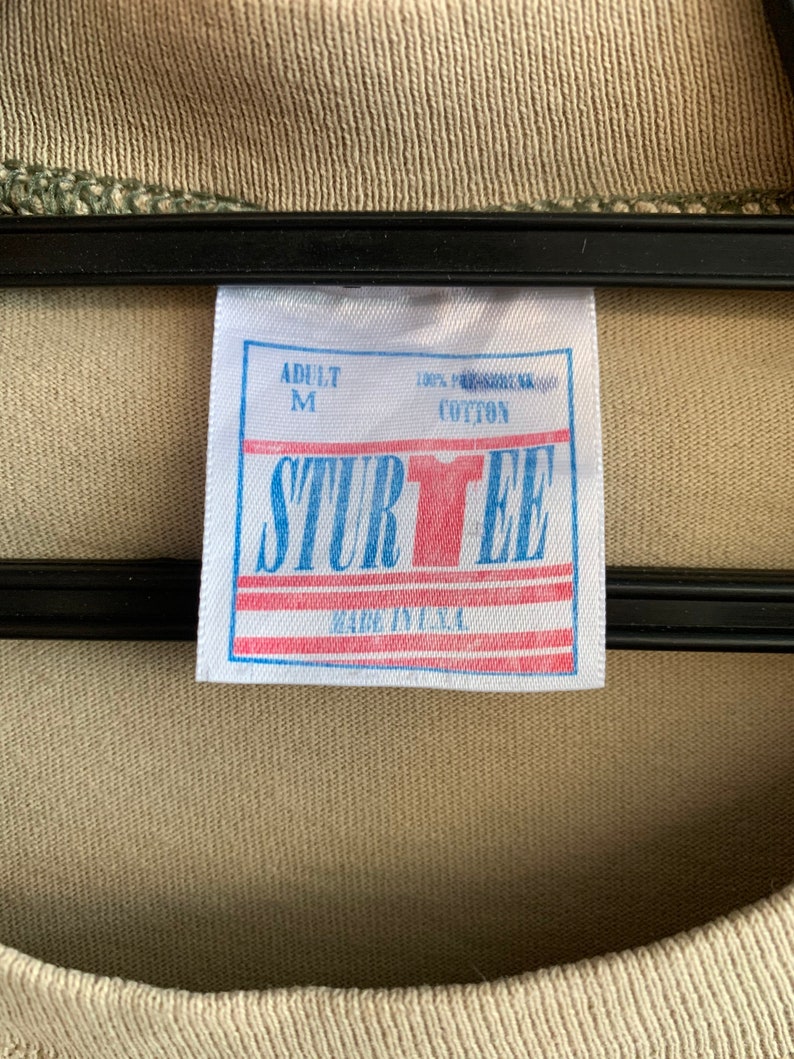 Puede incluir: Etiqueta blanca con texto azul que dice "Adult M 100% preshrunk cotton SturTee Made in U.S.A."