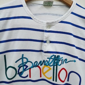 May include: White and blue striped t-shirt with a colorful embroidered Benetton logo. The tag says "Cotton, Baumwolle, Coton".