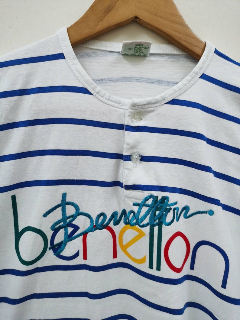 May include: White and blue striped t-shirt with a colorful embroidered Benetton logo. The tag says "Cotton, Baumwolle, Coton".