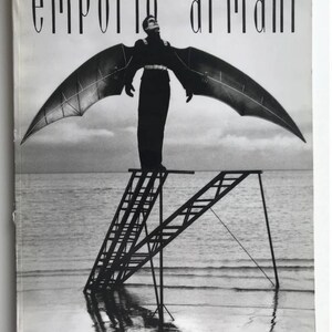 Puede incluir: Una fotograf&iacute;a en blanco y negro de un hombre con un atuendo negro y alas de pie en una escalera en el agua. El texto "emporio armani" est&aacute; en la parte superior de la imagen. El texto "MAGAZINE N&deg; 16" est&aacute; en la parte inferior izquierda de la imagen. El texto "ARMANI" est&aacute; en la parte inferior derecha de la imagen.