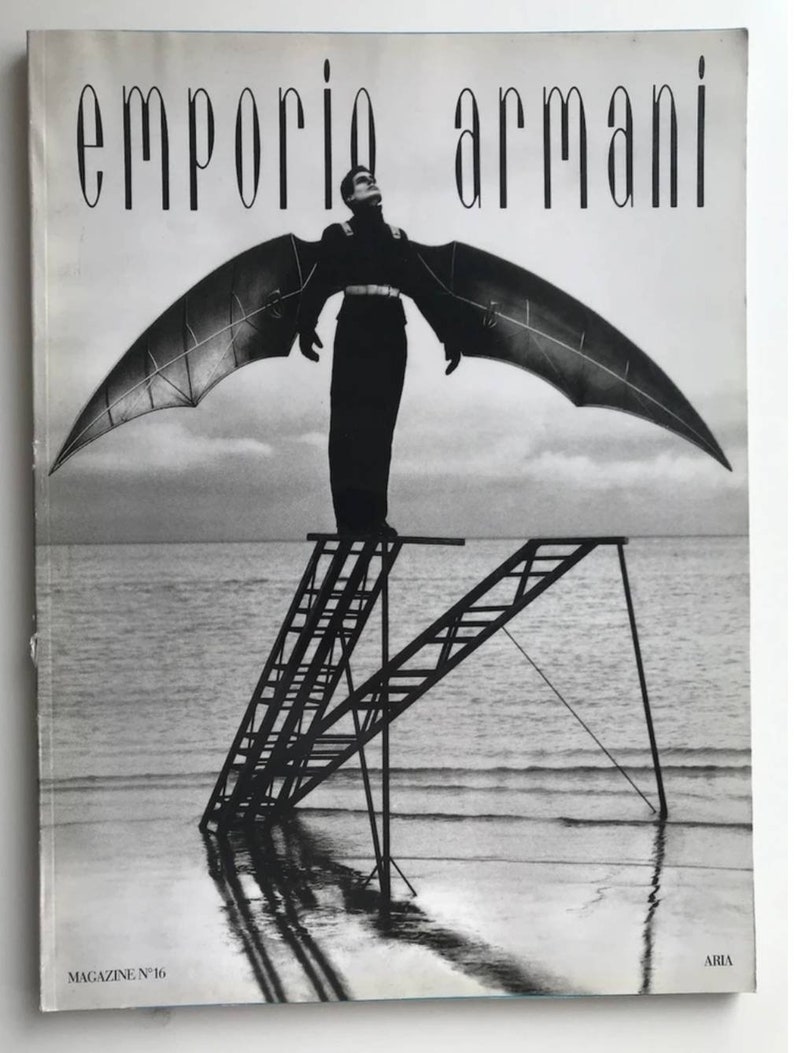 Puede incluir: Una fotograf&iacute;a en blanco y negro de un hombre con un atuendo negro y alas de pie en una escalera en el agua. El texto "emporio armani" est&aacute; en la parte superior de la imagen. El texto "MAGAZINE N&deg; 16" est&aacute; en la parte inferior izquierda de la imagen. El texto "ARMANI" est&aacute; en la parte inferior derecha de la imagen.