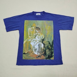 Puede incluir: Una camiseta azul con una pintura de una familia con un mono. La pintura está en un esquema de colores amarillo, verde y marrón. La camiseta tiene una etiqueta blanca con la palabra "Banana" en ella.