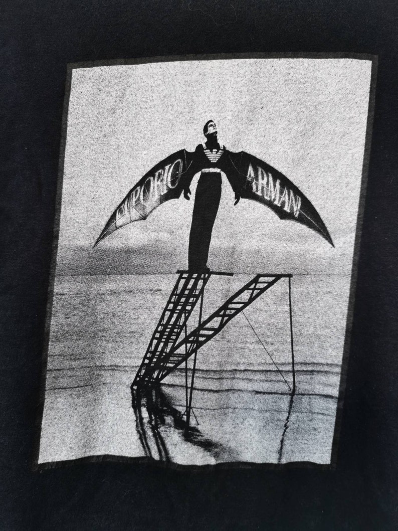 Puede incluir: Imagen en blanco y negro de una persona de pie en una escalera con alas que dicen "Emporio Armani". La persona est&aacute; de pie en una escalera que est&aacute; en una playa.