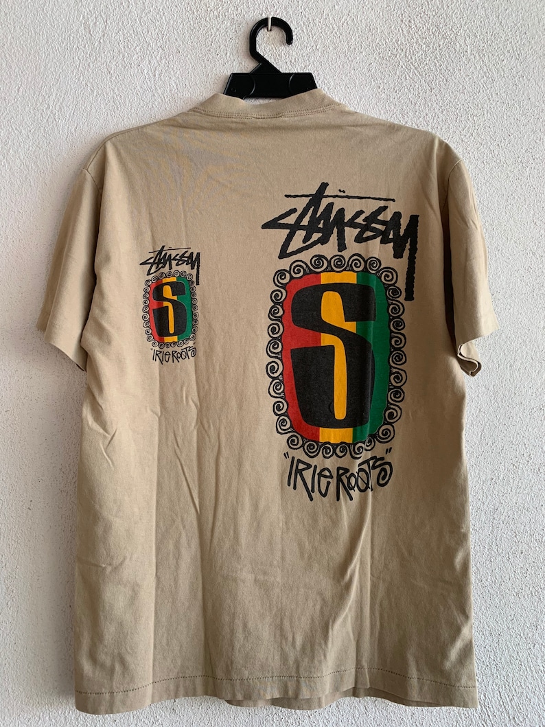 Puede incluir: Una camiseta de color marr&oacute;n claro con un gr&aacute;fico negro y verde de la letra "S" con las palabras "irie roots" impresas debajo. El gr&aacute;fico est&aacute; rodeado por un dise&ntilde;o de espiral negro y blanco. La palabra "Stussy" est&aacute; impresa en negro sobre el gr&aacute;fico.