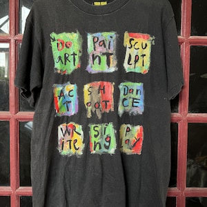 May include: Black t-shirt with a colorful graphic print of nine squares, each containing a different word related to art, such as "Do Art", "Paint", "Sculpt", "Act", "Shot", "Dance", "Write", "Sing", and "Play". The text "YOUR HEAR" is printed in white on a black tag at the top of the shirt.