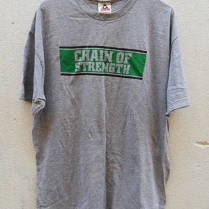 以下が含まれることがあります： グレーのTシャツに、緑と白の四角いグラフィックがプリントされています。グラフィックには「Chain of Strength」と書いてあります。