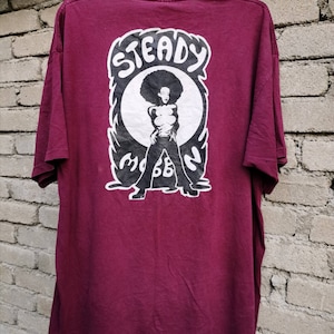 May include: A maroon t-shirt featuring a graphic of a woman with an afro and the words "Steady Mobbin" in a white and black design. The shirt is hanging on a black hanger against a brick wall.