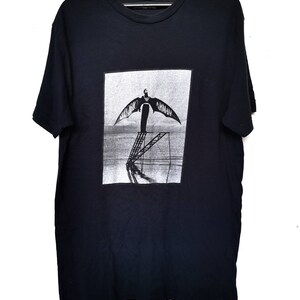 Puede incluir: Camiseta negra con un estampado gr&aacute;fico de una persona con alas de pie en una escalera. El texto "Emporio Armani" est&aacute; impreso en las alas.