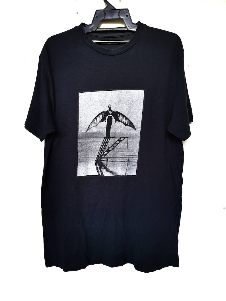 Puede incluir: Camiseta negra con un estampado gr&aacute;fico de una persona con alas de pie en una escalera. El texto "Emporio Armani" est&aacute; impreso en las alas.