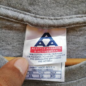 以下が含まれることがあります： グレーのTシャツタグに「Alstyle Apparel & Activewear」というテキストと「90% コットン/コトン/アルゴドン 10% ポリエステル/ポリエステル/ポリエステル プレシュランク/プレエンコジド プレレトレチ」というテキストと「Fabric Made in U.S.A. Assembled in Mexico」というテキストと「サイズ タイル タラ XL TG XG」というテキストが書かれています。