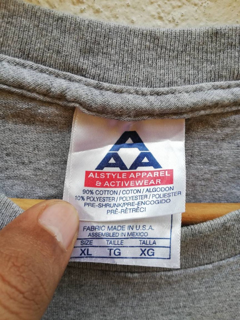 以下が含まれることがあります： グレーのTシャツタグに「Alstyle Apparel & Activewear」というテキストと「90% コットン/コトン/アルゴドン 10% ポリエステル/ポリエステル/ポリエステル プレシュランク/プレエンコジド プレレトレチ」というテキストと「Fabric Made in U.S.A. Assembled in Mexico」というテキストと「サイズ タイル タラ XL TG XG」というテキストが書かれています。