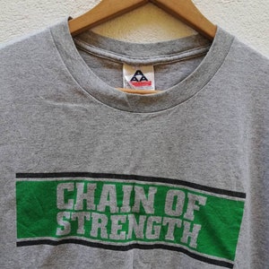 以下が含まれることがあります： グレーのTシャツに、緑と黒のグラフィックで「Chain of Strength」と書かれています。