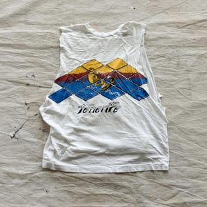May include: White sleeveless shirt with a colorful geometric design in blue, yellow, and red. The word "Tenerife" is printed in black below the graphic. The shirt is made of a soft, worn fabric.