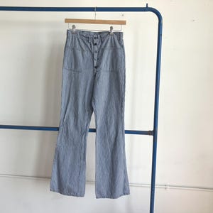 May include: High-waisted pants in a blue and white vertical pinstripe pattern. These pants feature a front button-up closure with visible dark buttons, two large patch pockets on the front, and a straight-leg cut hanging from a wooden hanger.