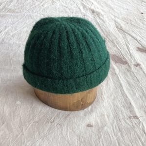 May include: A dark green, ribbed knit beanie hat with a folded brim. The hat is displayed on a wooden form against a white, textured background. The hat's color is a deep, forest green.