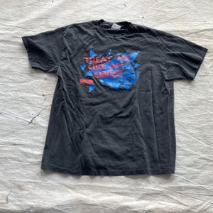 Könnte beinhalten: Dunkelgraues T-Shirt mit dem Text "TREAT ME LIKE AN OBJECT" in Rot und Blau. Die Grafik befindet sich auf einem blauen und roten Burst-Design. Das kurzärmelige T-Shirt ist wahrscheinlich aus Baumwolle.