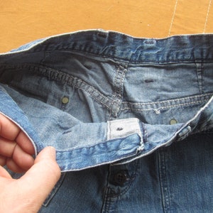 Op de afbeelding: Een blauwe spijkerbroek met een knoopsluiting en een tailleband. De broek is binnenstebuiten gekeerd om de stiksels en de voering te laten zien.