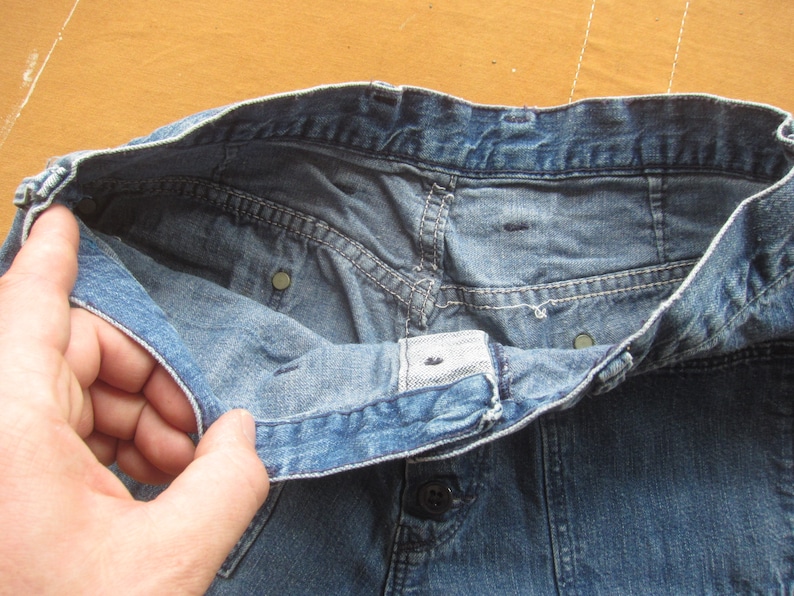 Op de afbeelding: Een blauwe spijkerbroek met een knoopsluiting en een tailleband. De broek is binnenstebuiten gekeerd om de stiksels en de voering te laten zien.