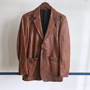 May include: A brown leather blazer with decorative stitching on the lapels and pockets, hanging on a wooden hanger. The blazer has a single-button closure and a black lining. The jacket is displayed against a white background.