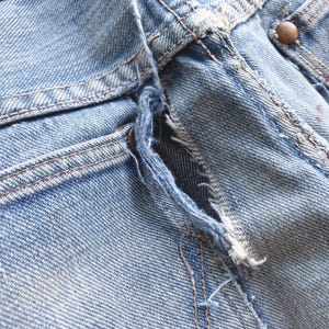 Peut inclure: Gros plan sur un jean en denim bleu clair, montrant une zone ab&icirc;m&eacute;e pr&egrave;s de la braguette. Le tissu est us&eacute; et effiloch&eacute;, avec des coutures visibles et un bouton cuivr&eacute;. Le jean a une conception classique &agrave; cinq poches.