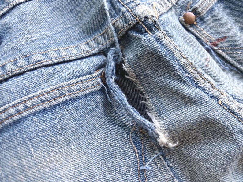 Peut inclure: Gros plan sur un jean en denim bleu clair, montrant une zone ab&icirc;m&eacute;e pr&egrave;s de la braguette. Le tissu est us&eacute; et effiloch&eacute;, avec des coutures visibles et un bouton cuivr&eacute;. Le jean a une conception classique &agrave; cinq poches.