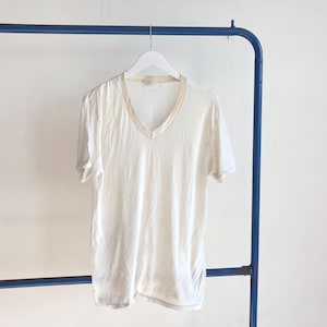 May include: A short-sleeved V-neck t-shirt in an off-white or cream color. The shirt features a slightly darker, beige-toned trim around the neckline and is displayed on a white plastic hanger against a plain, light-colored background.