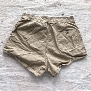May include: Khaki-colored shorts with a high waist and a single back pocket. The shorts have a button closure and belt loops. The fabric appears to be a soft, woven material. The shorts are laid flat on a textured, off-white surface.