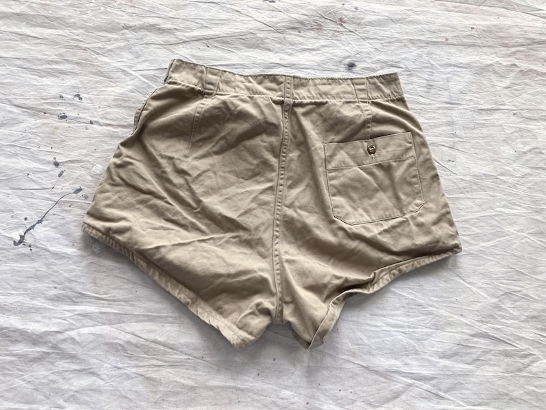 May include: Khaki-colored shorts with a high waist and a single back pocket. The shorts have a button closure and belt loops. The fabric appears to be a soft, woven material. The shorts are laid flat on a textured, off-white surface.