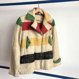 May include: A vintage-style jacket with a cream-colored base and horizontal stripes in red, yellow, and black. The jacket features a full zipper, two front pockets, and green accents on the collar and pockets. It is hanging on a wooden hanger.