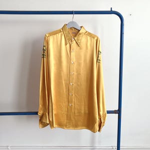 Puede incluir: Una camisa dorada de manga larga con cuello clásico y abotonadura frontal. La camisa tiene detalles bordados en verde en las mangas. La camisa está colgada en una percha de metal azul.
