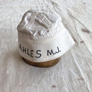 May include: A white bucket hat with the text "AHLES M.J." in black lettering. The hat is sitting on a wooden form. The hat has a brim and is made of a soft fabric.